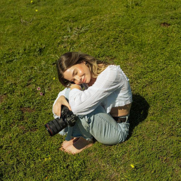 Young woman sitting barefoot on grass holding a camera, eyes closed peacefully.