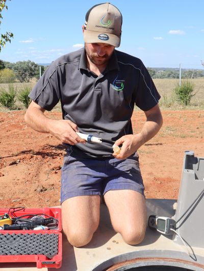 Man in uniform kneeling outdoors, working with a handheld tool and electronic equipment.