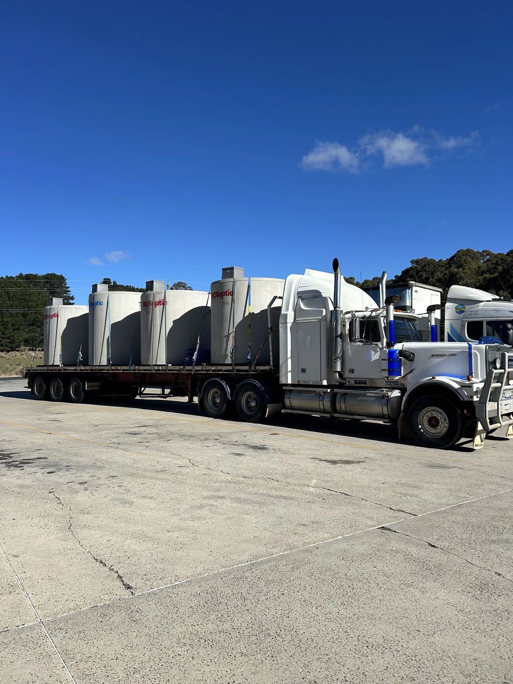 Large white truck transporting tall septic tanks on a sunny day.