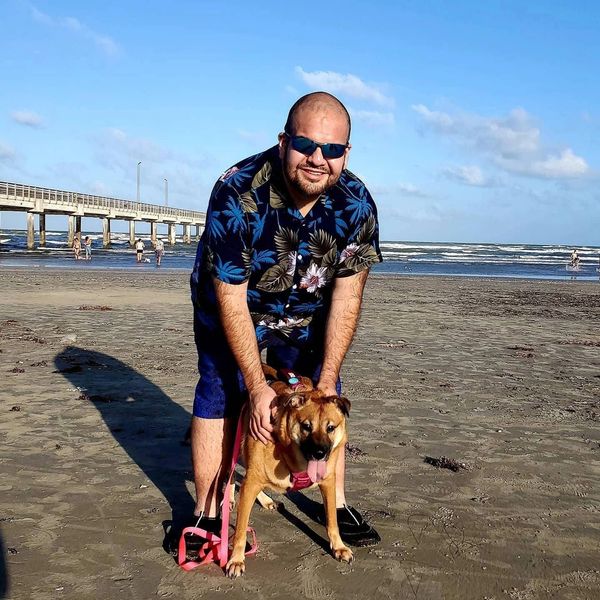 Man in floral shirt with dog on a beach.