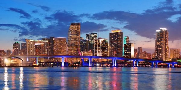 City skyline at dusk with illuminated buildings and a blue-lit bridge over water.