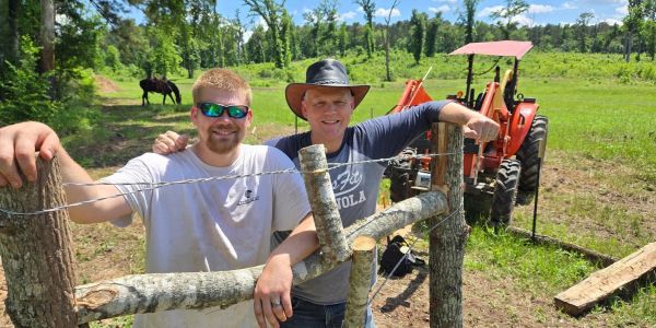 Two men standing by a wooden fence with a tractor in the background.
