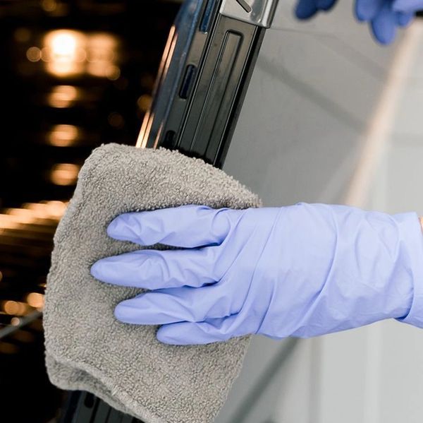 A gloved hand cleaning an oven door with a cloth.
