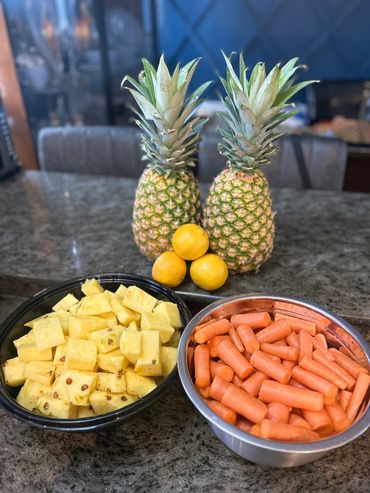Two pineapples, three lemons, cubed pineapples, and baby carrots on a kitchen counter.