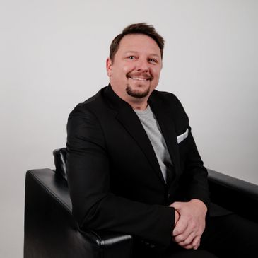 Man in black suit sitting on a black chair, smiling.