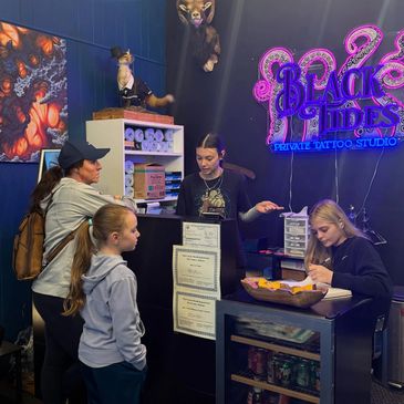 Customers and staff at a tattoo studio reception desk with neon sign.