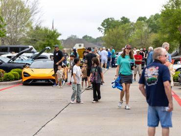 People walk and admire cars at an outdoor car show on a cloudy day.
