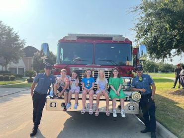 Children sitting on a fire truck bumper with two firefighters standing beside them.