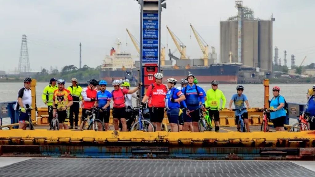 Cyclists gathered near a control tower by an industrial port with cranes and ships.