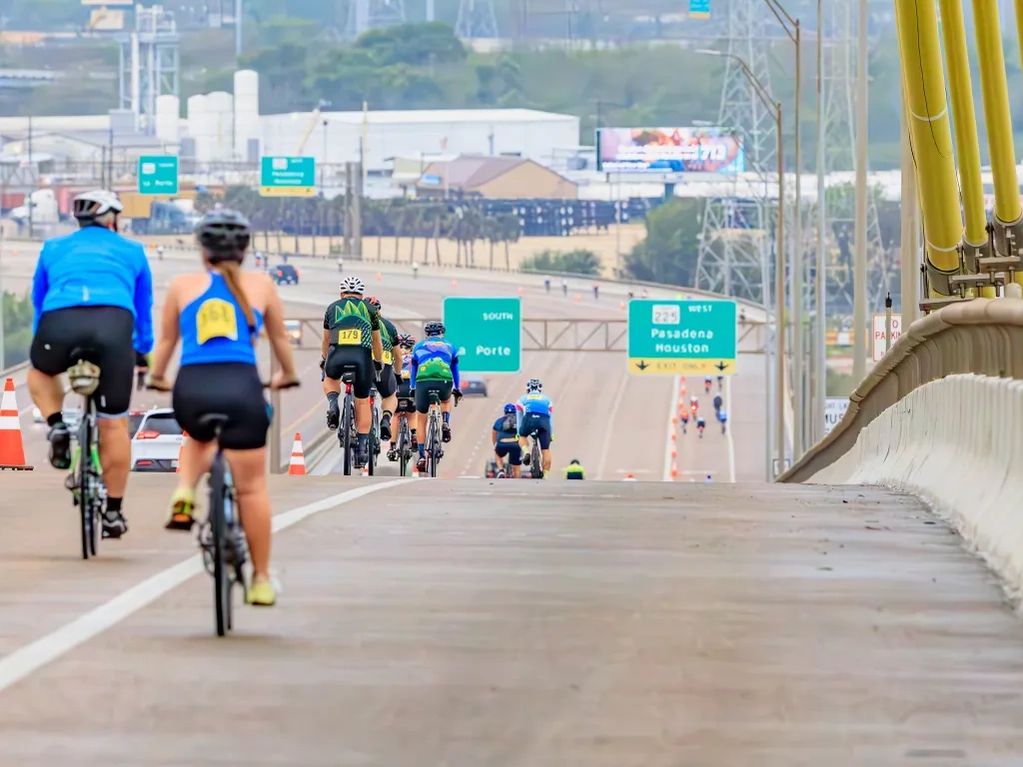 Cyclists riding on a highway with signs for Pasadena and Houston.