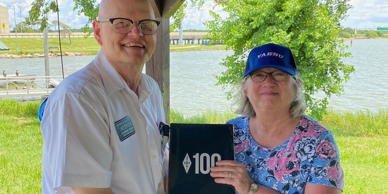 Two smiling people holding a black book with '100' on the cover by a lakeside.