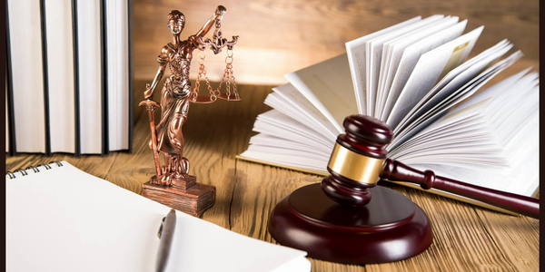 A wooden desk with a notebook, a pen, a gavel, a small statue of Lady Justice, and various books.
