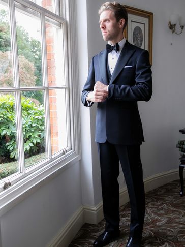 Man in a tuxedo adjusting his cuff by a window, looking thoughtfully outside.