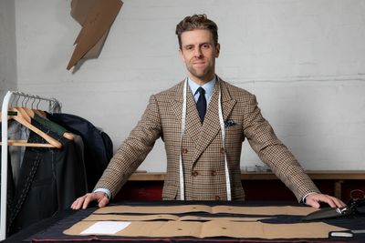 Tailor in a checkered blazer with measuring tape, standing by a table with fabric and patterns.