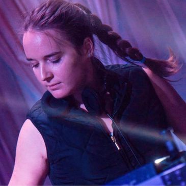 A focused female DJ with braided hair performing at a dimly lit event.