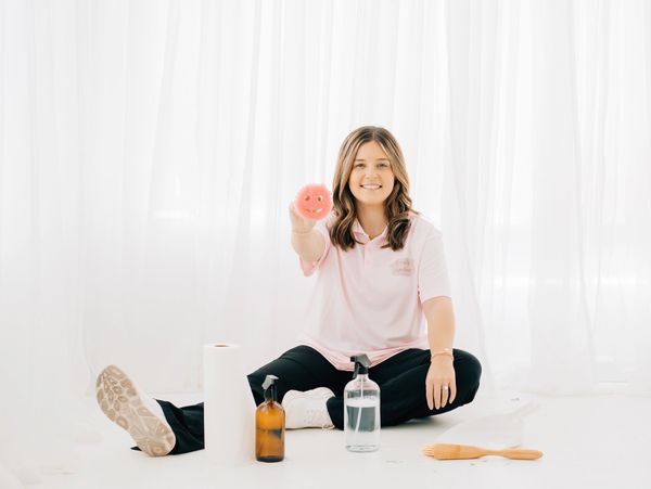 Smiling woman holding a pink smiley face sponge, surrounded by cleaning supplies.