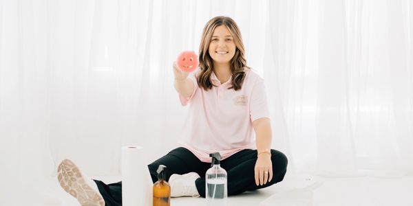 Smiling woman sitting with cleaning supplies and holding a smiley face sponge.