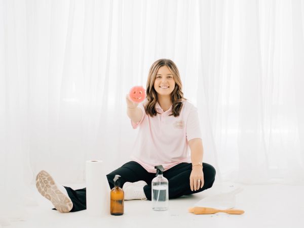 Smiling woman holding a pink smiley face scrubber, surrounded by cleaning supplies.