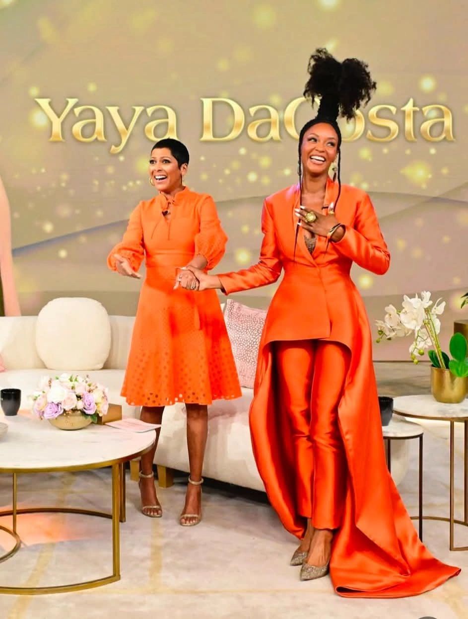 Two women in vibrant orange outfits smiling and holding hands on a talk show set.
