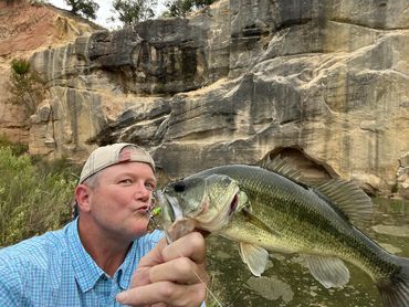 STOCKED POND, FISHERMAN POSES WITH CATCH IN FRONT OF A CLIFF FACE
