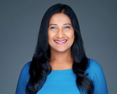 Smiling woman with long black hair wearing a blue top against a gray background.