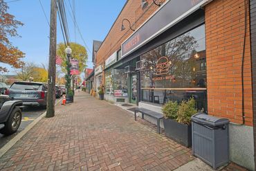 Street view of a brick sidewalk with shops and parked cars.