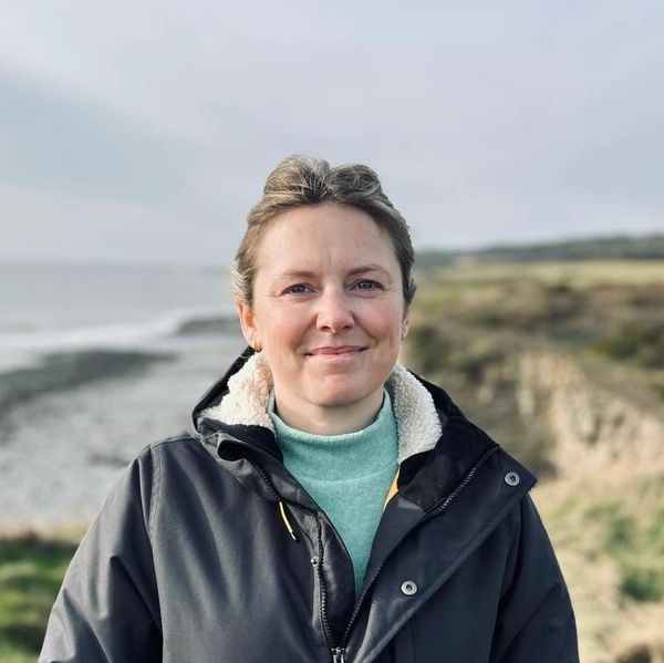 Smiling woman in a warm jacket stands outdoors near a coastline on a cloudy day.