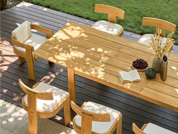 Outdoor wooden dining table with cushioned chairs on a deck under dappled sunlight.