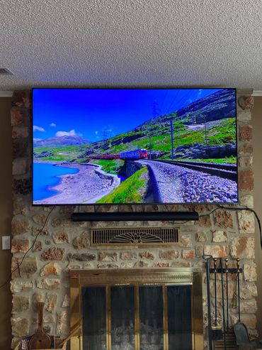 A TV mounted above a stone fireplace showing a scenic train traveling by a lake and green hills.