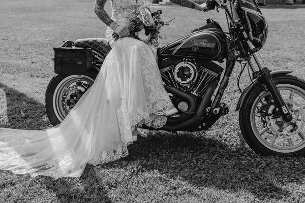Bride in a lace wedding dress sitting on a Harley-Davidson motorcycle, holding a bouquet.