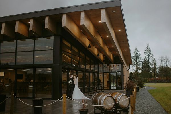 A newlywed couple embraces outside a modern building with wooden accents and barrels nearby.