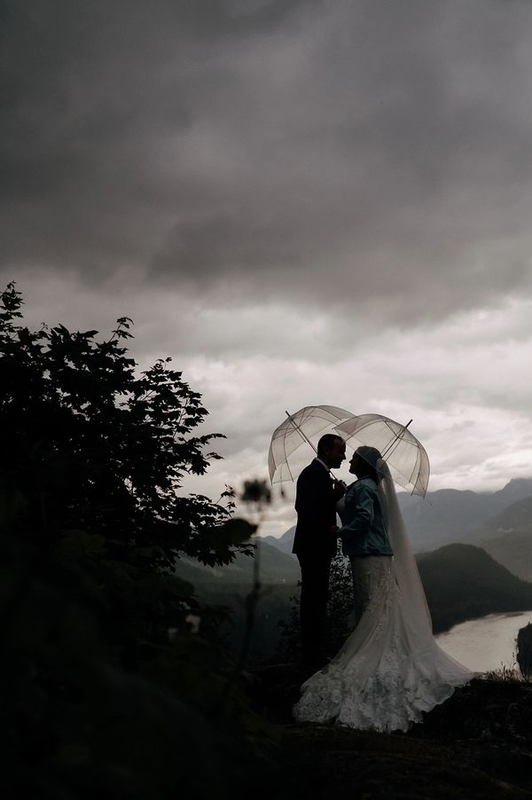 Silhouetted couple in wedding attire share a moment under umbrellas against a moody sky.