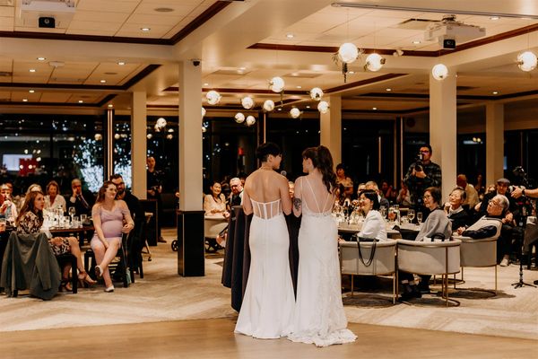 Two brides in elegant white gowns speak to guests at a wedding reception.