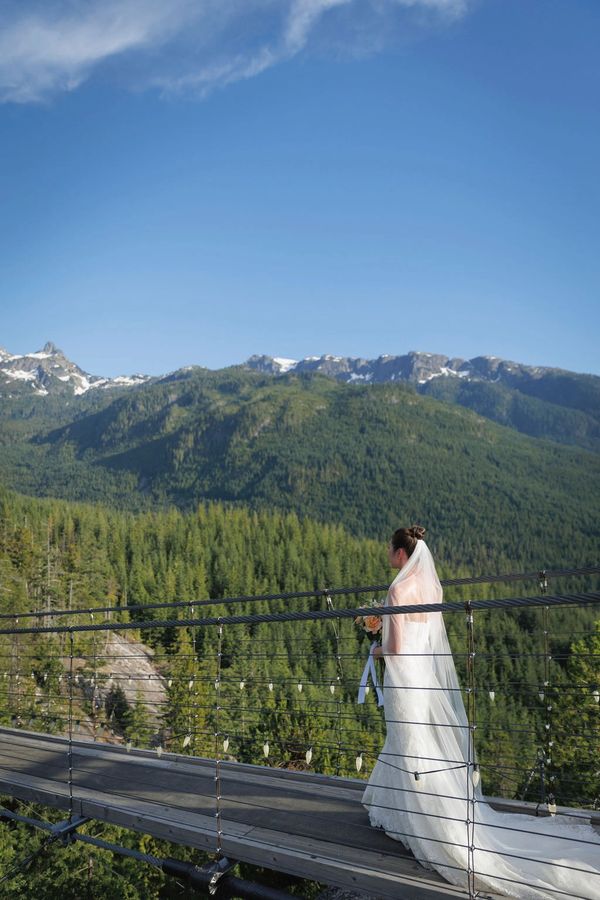 Bride in white gown stands on a suspension bridge overlooking forested mountains.