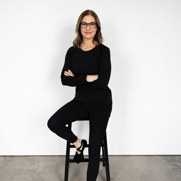 Confident woman in black outfit sitting on a stool against a white background.