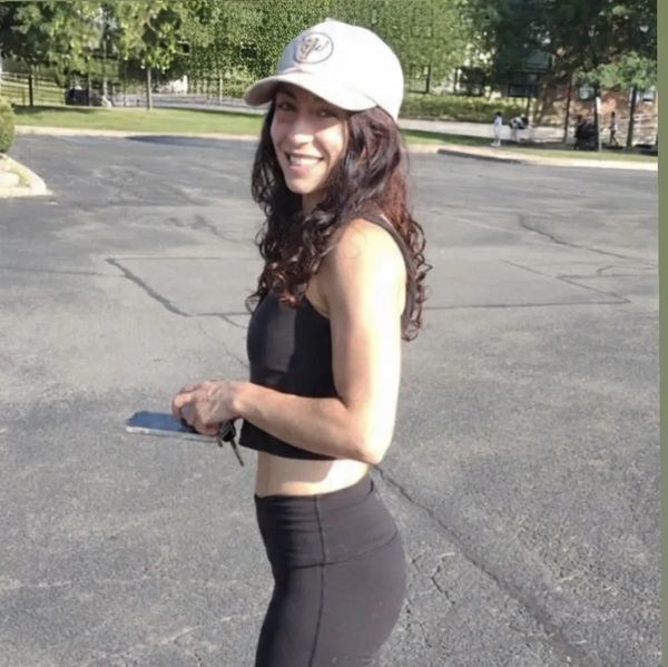 Woman in black workout outfit smiling outdoors with a white cap.