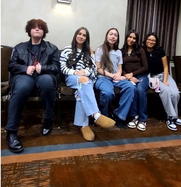 Five young people sitting on chairs indoors.