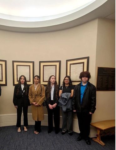 Five young people standing indoors near framed documents.
