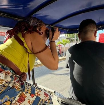 A woman in colorful pants takes photos while riding a tricycle taxi driven by a man.