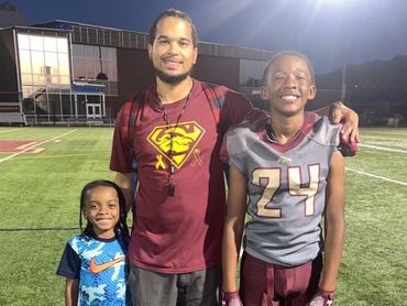 Three smiling males on a football field at night, one in football gear.