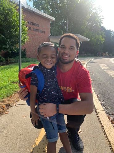 A man and a child smiling outside a school on a sunny day.