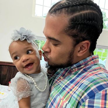 A man lovingly kisses a smiling baby girl dressed in white with a pearl necklace.