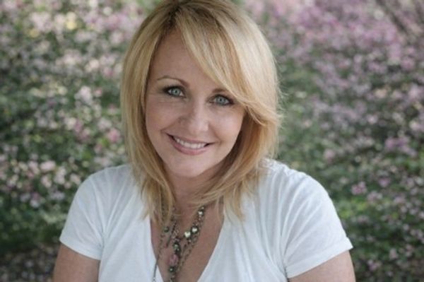 Smiling woman with blonde hair outdoors against a flowering bush.