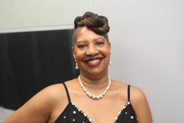 Woman smiling confidently in a black dress with pearls.