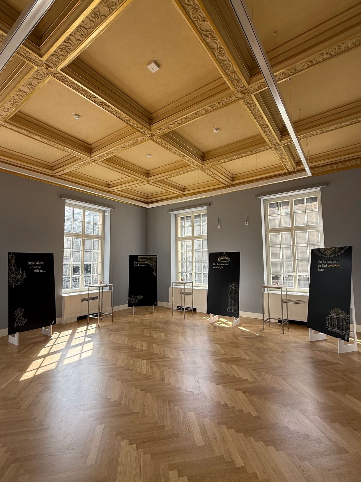 Elegant exhibition room with ornate ceiling and black display boards near large windows.