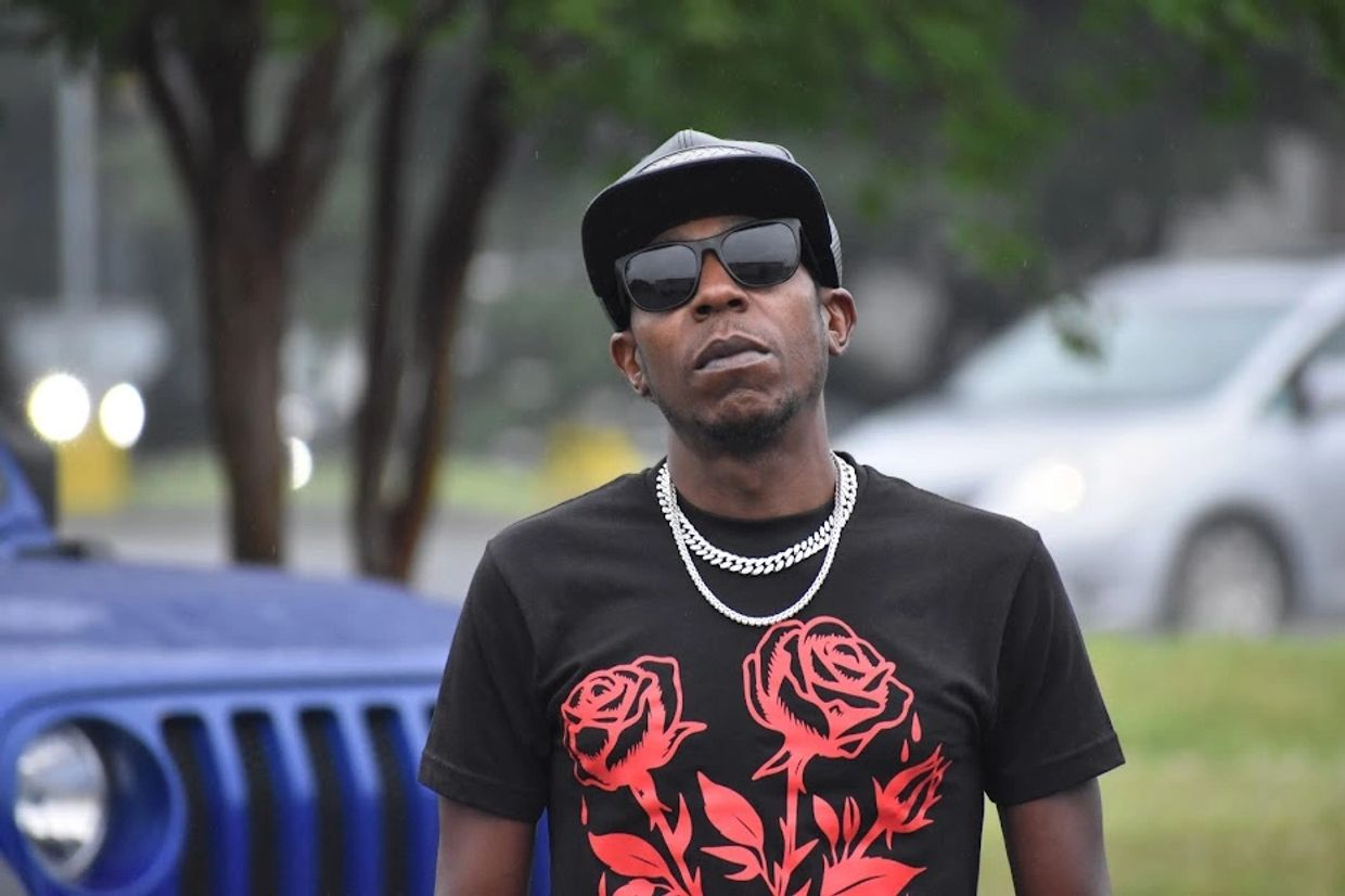 Man in sunglasses and black cap with rose shirt standing outdoors.
