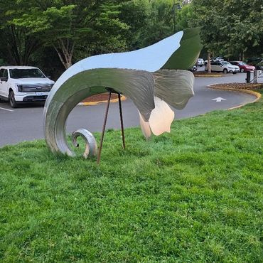 Curved metal sculpture resembling a wave or leaf on a grassy area near a parking lot.