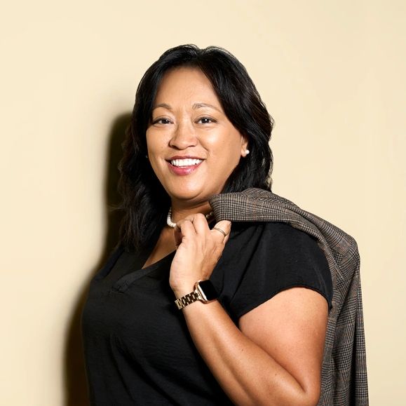 Confident woman smiling, holding a jacket over her shoulder against a beige background.