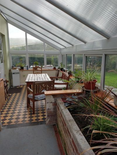 A sunlit conservatory with wooden furniture and numerous potted plants.