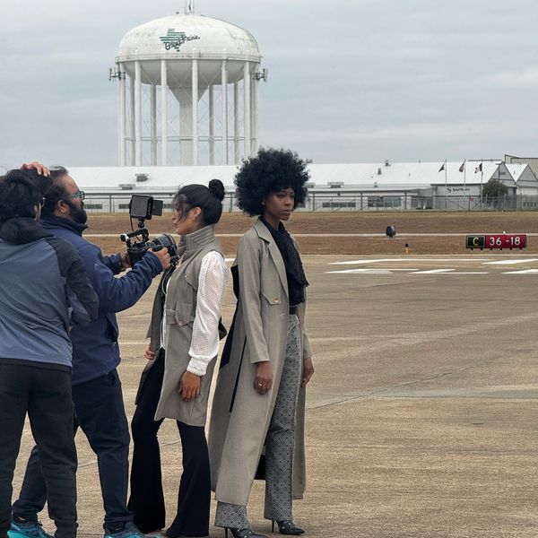 Two stylish women posing for a video shoot on an airfield with a water tower in the background.